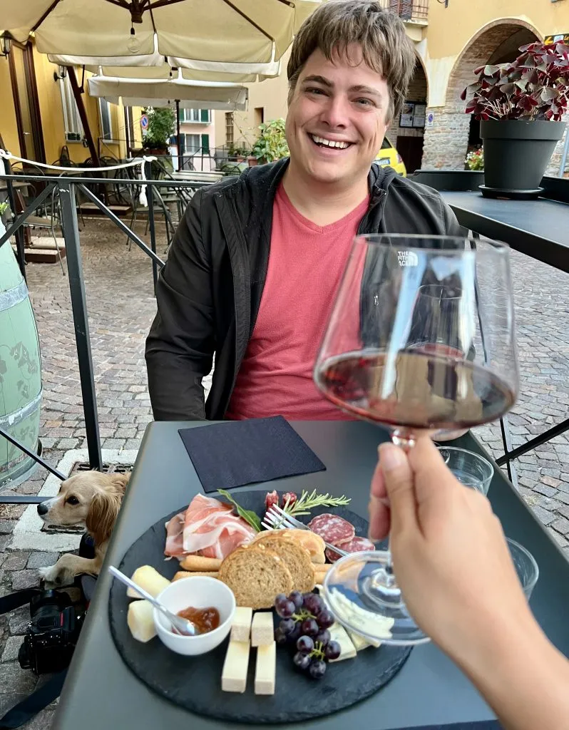 jeremy storm at a wine bar in barolo italy with a cheese plate on the table in front of him and a wine glass in the foreground