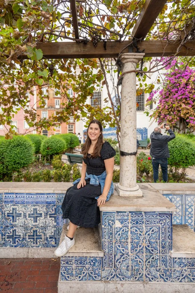 kate storm in a black dress sitting on a ledge at miradouro de santa luzia, one of the best places to visit in lisbon in 3 days