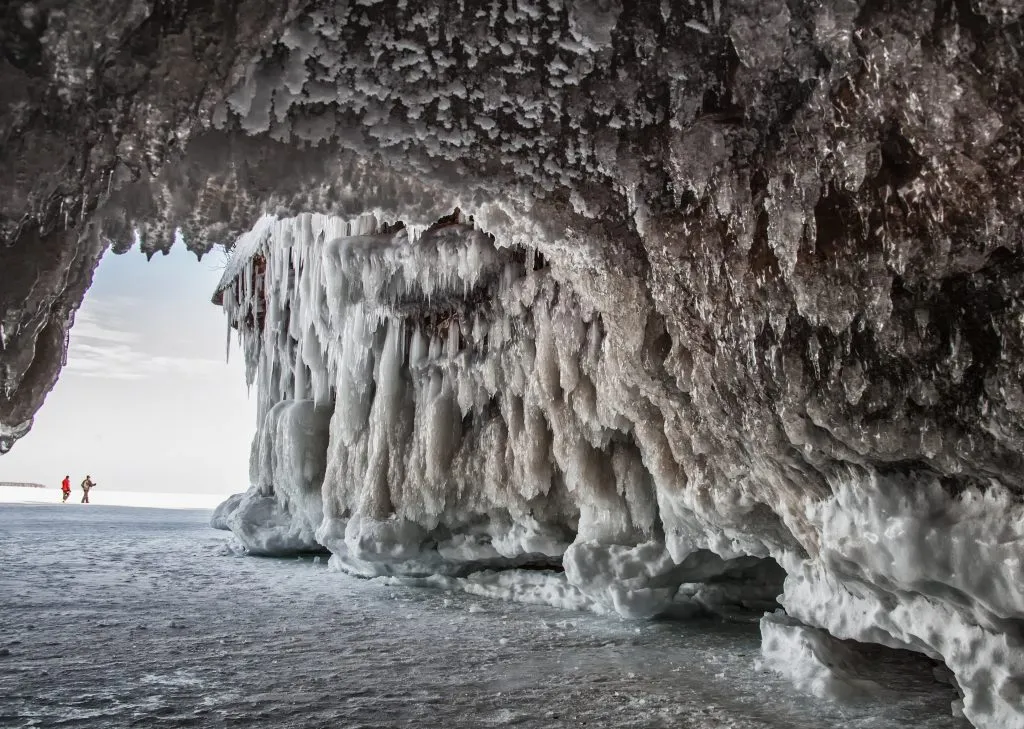 two people hiking in front of an ice cave at apostle islands, a fun travel destination in the usa in winter