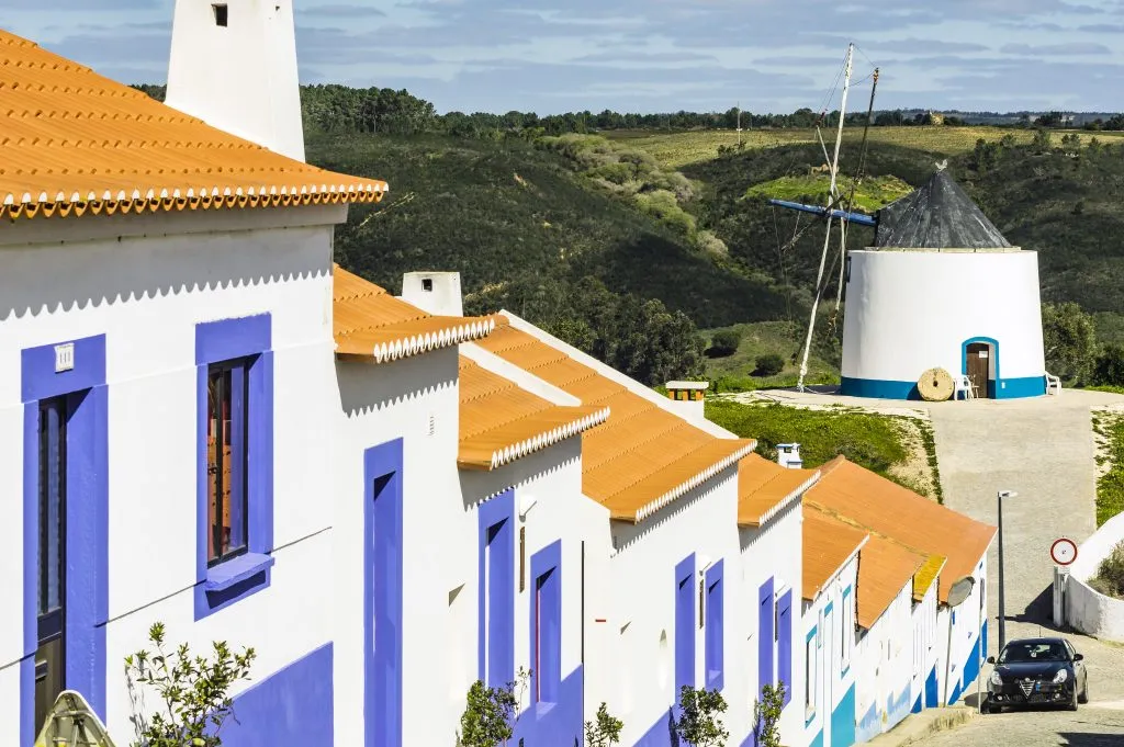 white buildings with purple trim and a windmill in the background in odeceixe portugal