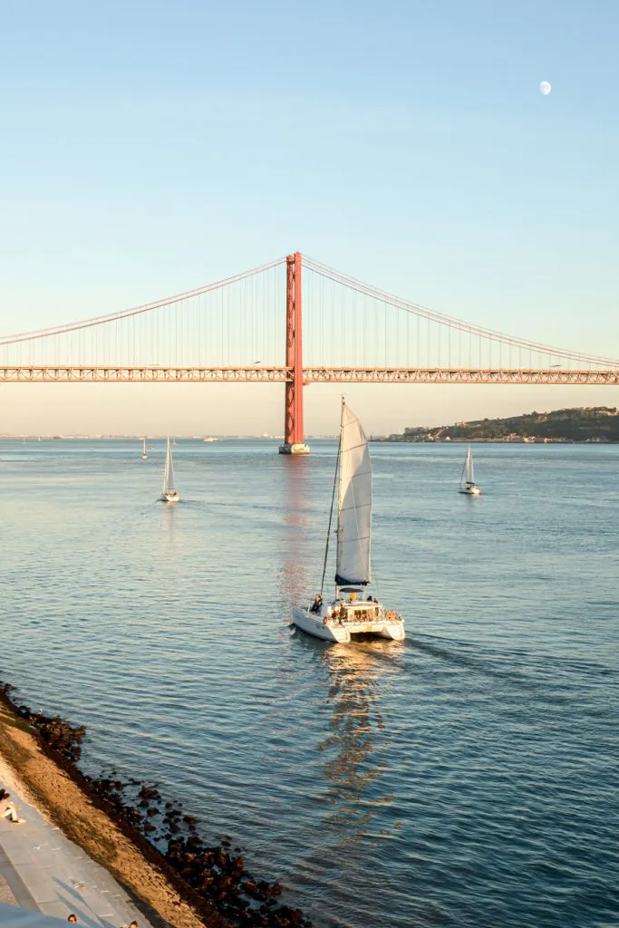 sailboats moving along the tagus river at sunset with ponte 25 de abril in the background, one of the best things to do in lisbon belem