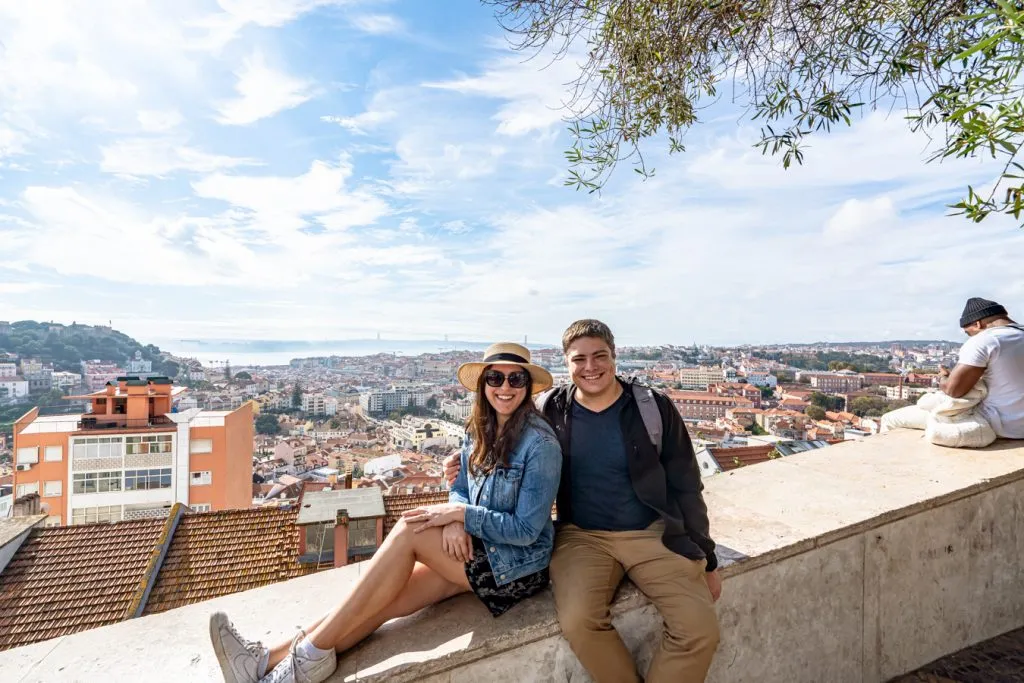 kate storm and jeremy storm sitting on a wall overlooking lisbon portugal