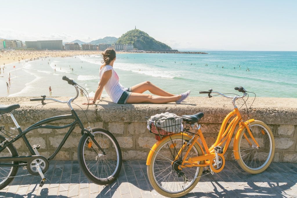 young woman sitting on a stone wall overlooking the beach in san sebastaian spain with bikes in the foreground