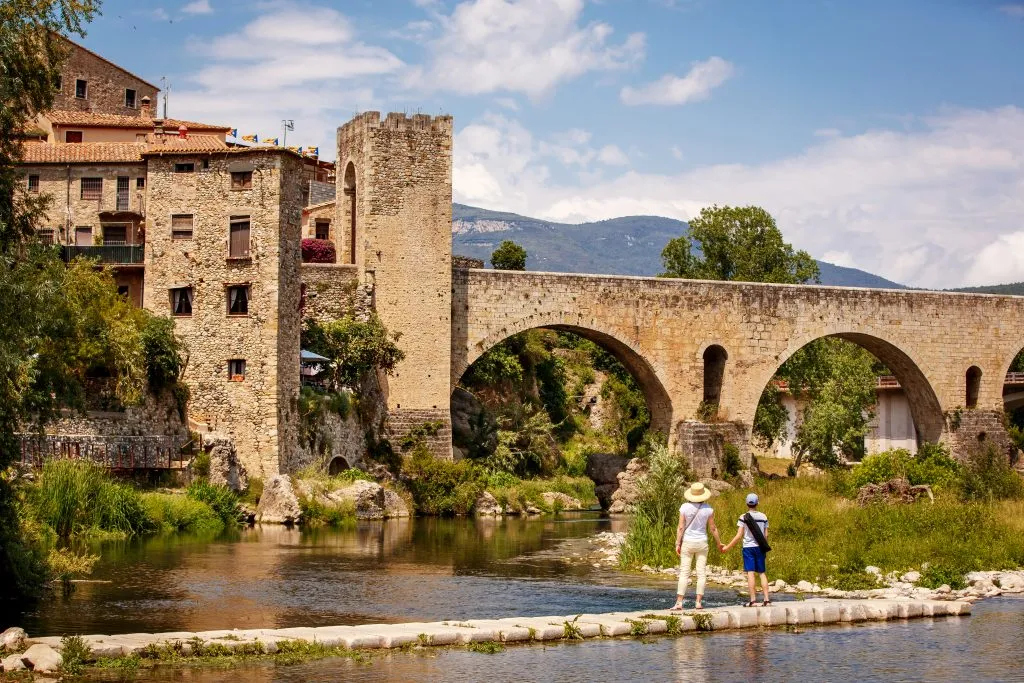 two people standing along a river looking up at a stone bridge in besalu, one of barcelona best day trips