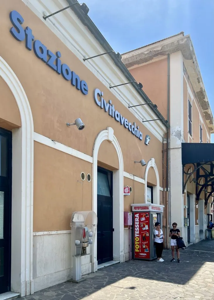 front facade of the civitavecchia italy train station, yellow building with "stazione civitavecchia" written in blue letters on the side