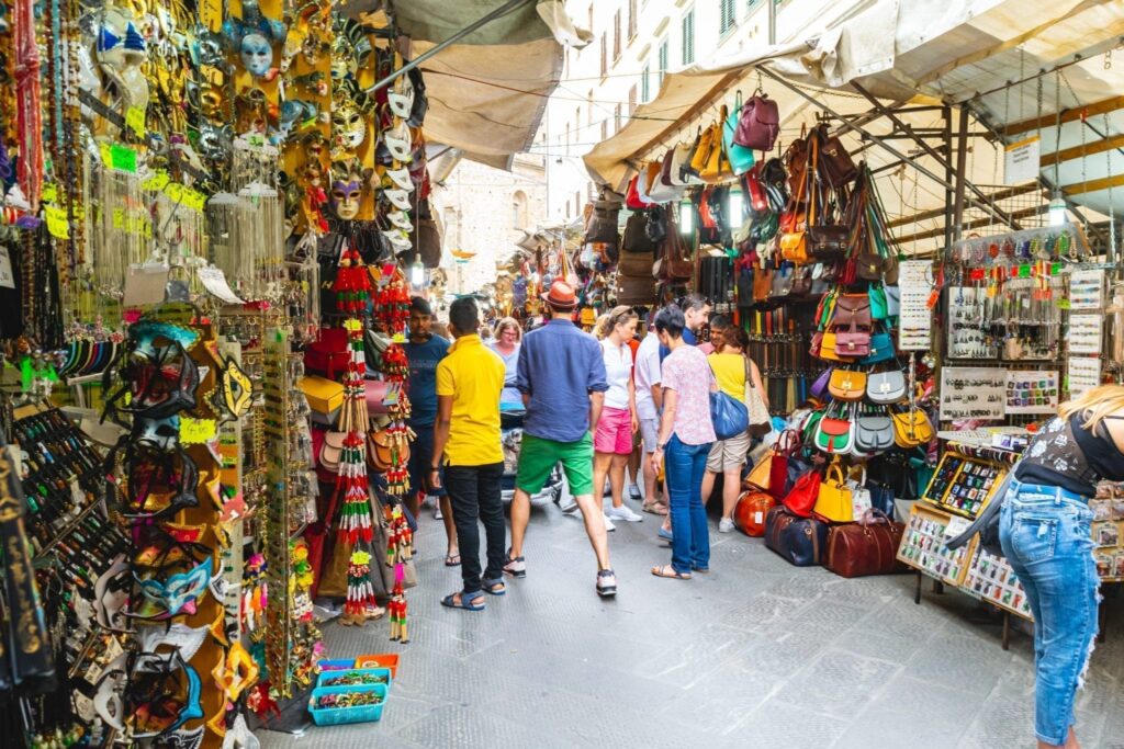 tourists walking through mercato san lorenzo souvenir shopping in florence italy with souvenirs displayed on both sides of the street