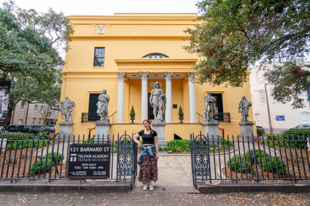 kate storm standing in front of the telfair academy yellow building with statues in front of it during a long savannah weekend getaway itinerary